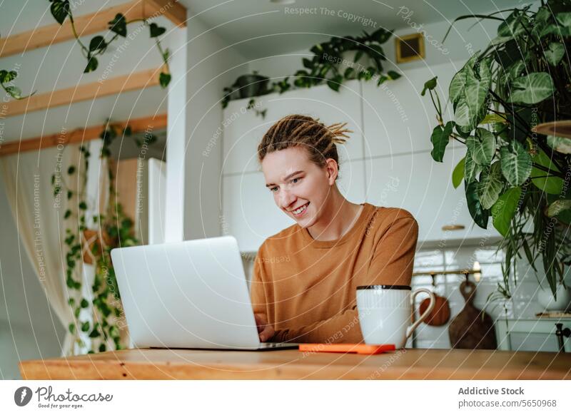 Glückliche junge Frau arbeitet am Laptop am Küchentisch mit Smartphone und Kaffeetasse im Tageslicht Kabinett Tisch zu Hause Rastalocken Apparatur Gerät Mobile