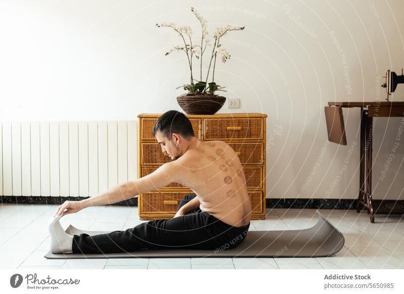 Konzentrierter junger Mann sitzt auf einer Matte und streckt sein Bein beim Training zu Hause Dehnung Rehabilitation heimwärts üben Yoga meditieren Sitzung