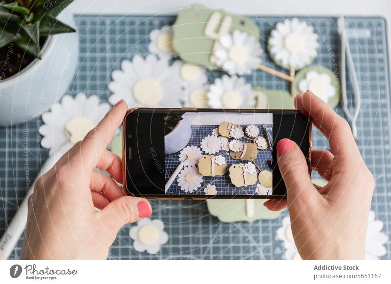 Frau, die durch einen Smartphone-Bildschirm eine kreative Ausstellung von handgemachten Papieranhängern und Gänseblümchen einrahmt Hand Fotografie Basteln Tag