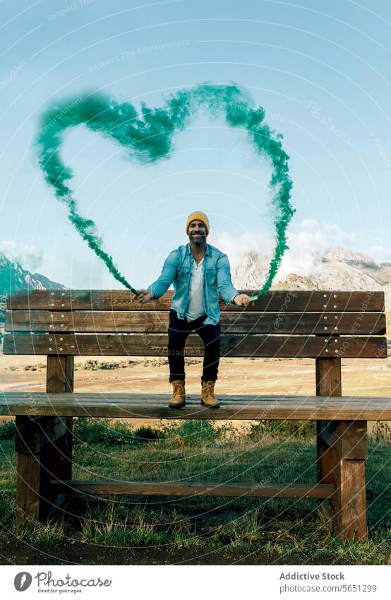 Mann mit herzförmigem Rauch, der eine skurrile Szene erzeugt Bank Herz grün Freude Fröhlichkeit malerisch Berge Landschaft Natur Liebe Konzept kreativ
