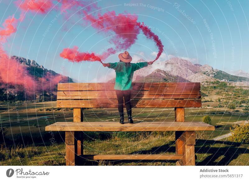 Fröhliche Frau steht auf einer großen Bank mit farbigem Rauch vor einem Berghintergrund Person Rauchfackel farbiger Rauch rot rosa Berge u. Gebirge Landschaft