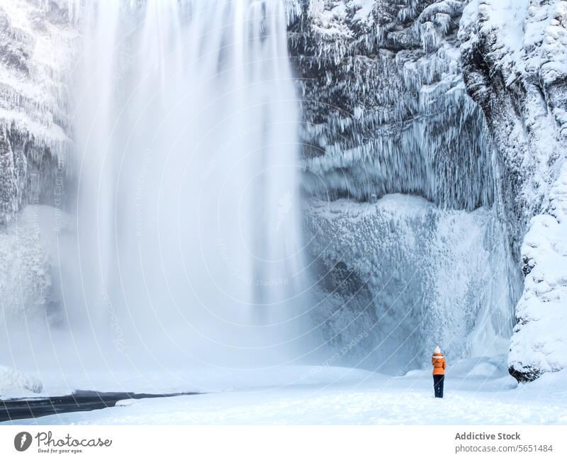 Eine anonyme Person in einer orangefarbenen Jacke steht ehrfürchtig vor dem majestätischen, eisigen Skógafoss-Wasserfall in Island Skogafoss Winter Eis Schnee