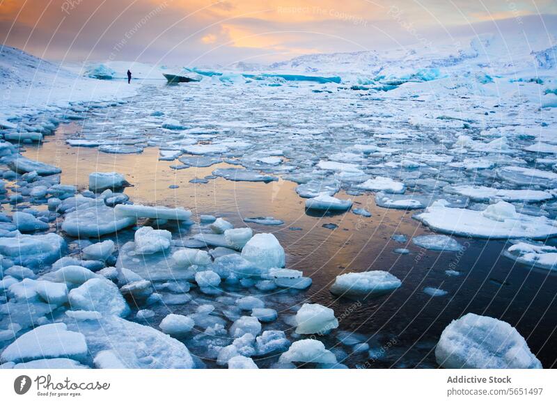 Eine Person steht am Rande einer mit Eisbrocken gefüllten Gletscherlagune unter einem pastellfarbenen Sonnenuntergangshimmel in Island Himmel Pastell Winter