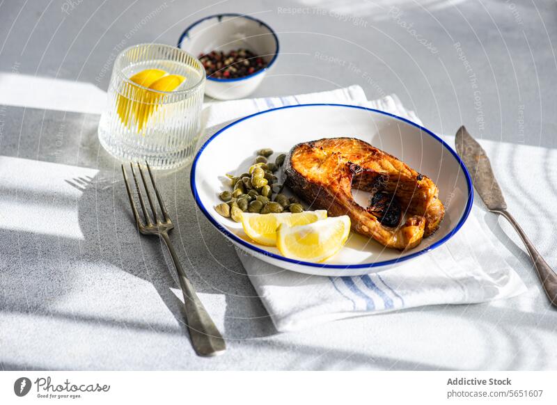 Von oben gegrilltes Forellensteak auf einem Teller mit Kapern und Zitrone auf dem Tisch neben einem Glas Wasser und Besteck Steak weiß blau Rand Garnierung