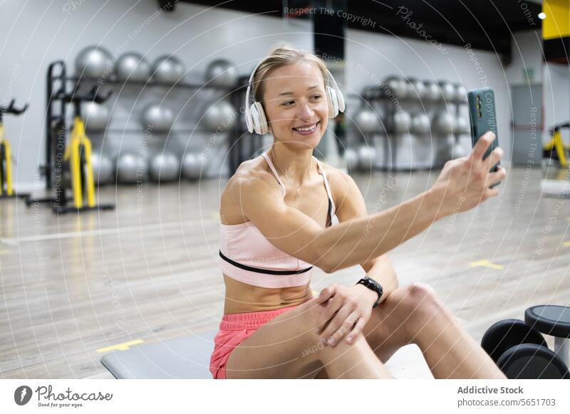 Lächelnde Frau nimmt Selfie im Fitnessstudio während des Trainings Sportbekleidung Kopfhörer Smartphone Übung Fröhlichkeit Technik & Technologie Lifestyle