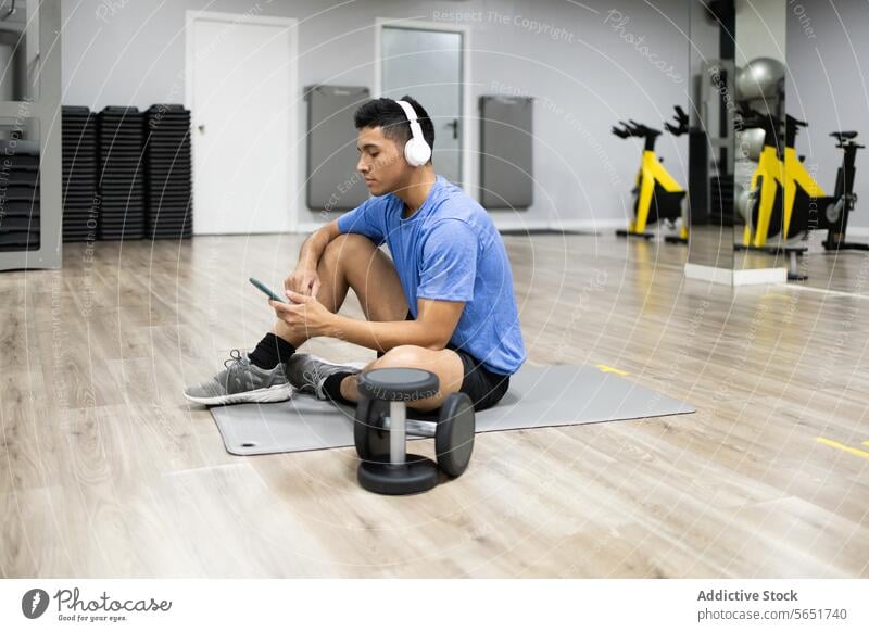 Junger Mann benutzt sein Smartphone in der Sportpause Fitnessstudio Übung Gerät Pause Kopfhörer Sitzen Unterlage fokussiert jung Erwachsener