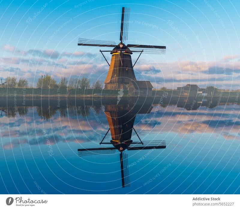 Einsame Windmühle, die sich im stillen Wasser eines holländischen Kanals in der Morgendämmerung bei strahlend blauem Himmel spiegelt Reflexion & Spiegelung