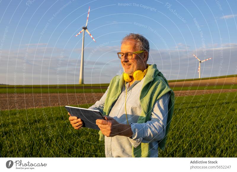 Fröhlicher älterer Mann mit Kopfhörern und digitalem Tablet im Windpark digitales Tablett Glück Windmühle Bauernhof Blauer Himmel Senior benutzend Ackerbau