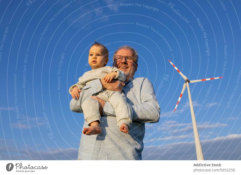 Von unten lächelnder älterer Mann, der ein Baby hält, mit einer Windkraftanlage im Hintergrund unter einem klaren blauen Himmel Großvater Enkel Blauer Himmel