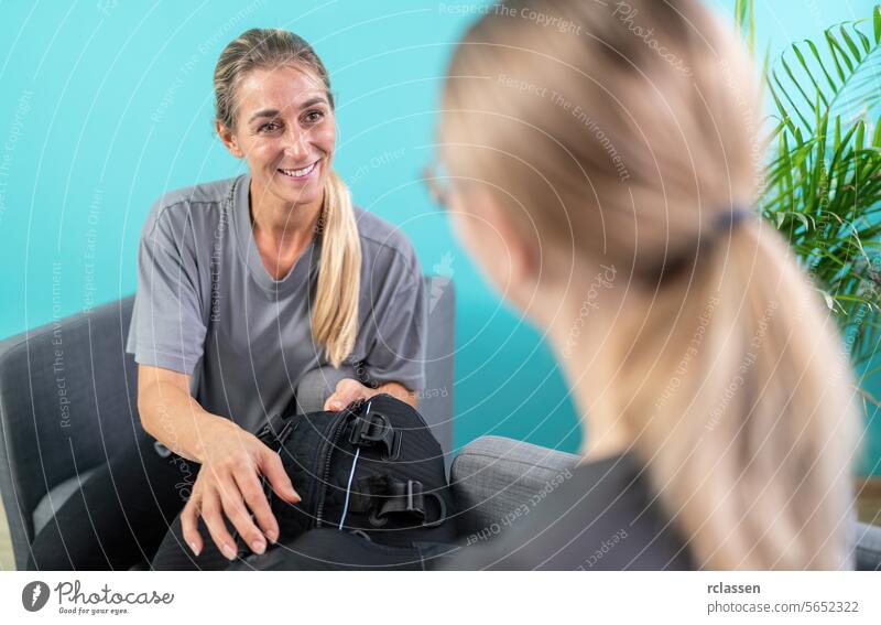 Frau hält einen EMS-Schulungsanzug in der Hand, während sie mit einem Ausbilder in einem Beratungsraum spricht ems-trainingsanzug Trainerin sprechend