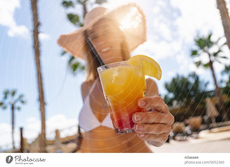 Nahaufnahme eines bunten Cocktails in der Hand einer Frau im Bikini, mit einem Strohhut und Palmen in einem karibischen Inselhotel Party Mallorca Hotel