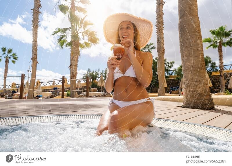 Frau in weißem Bikini nippt an einem Cocktail in einem Whirlpool, umgeben von Palmen auf einer Karibikinsel Hotel Pool trinken Spaß Resort jubelt Hut sonnig