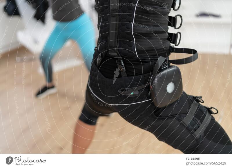 Zwei Frauen machen Kniebeugen in einem Fitnessstudio, eine trägt einen EMS-Trainingsanzug mit Akkupack Elektro Anregung neuromuskulär elektrisch muskulös