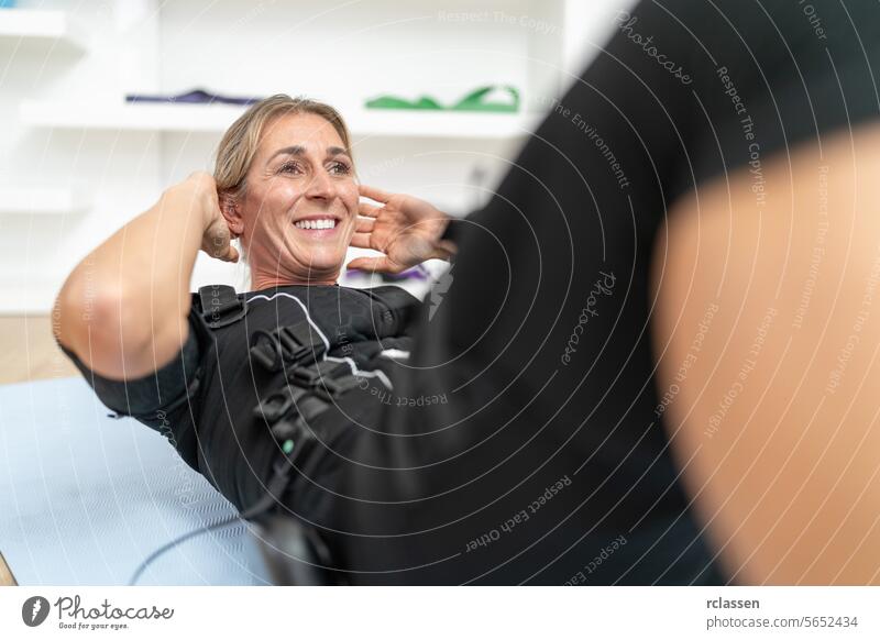Frau lächelt, während sie in einem EMS-Trainingsanzug in einem Fitnessstudio Sit-ups macht neuromuskulär ems-trainingsanzug Lächeln Übung persönliche Ausbildung