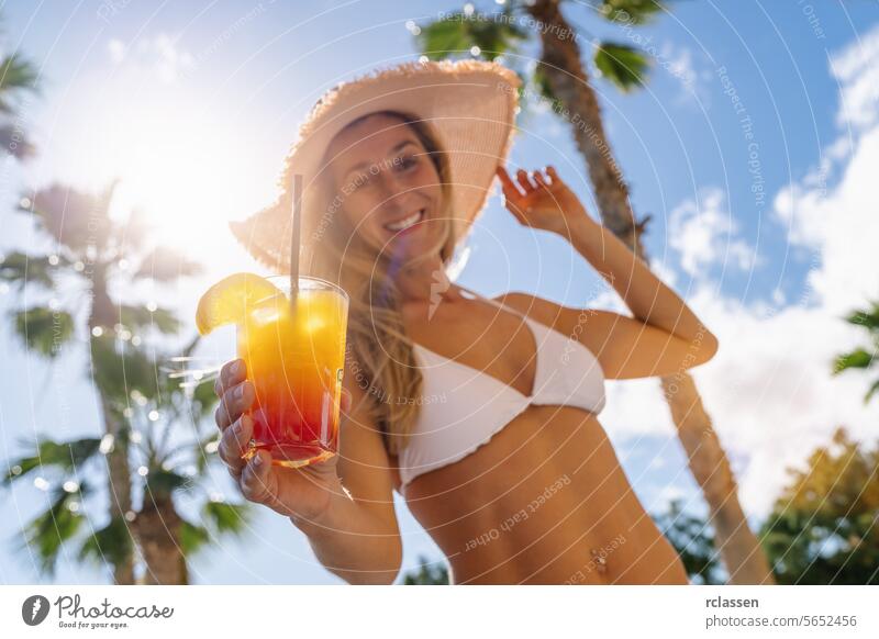 Lächelnde Frau in weißem Bikini und Strohhut mit einem tropischen Cocktail in der Hand, Palmen, sonniger Himmel auf einer Karibikinsel Party lächelnde Frau