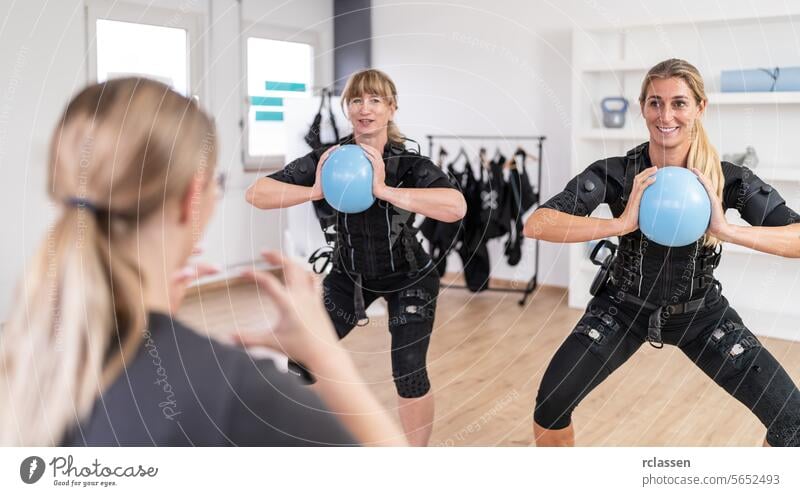 Frauen trainieren mit blauen Gymnastikbällen vor einem Trainer in einer Trainingseinheit und tragen EMS-Trainingsanzüge in einem EMS-Studio. schwarzer Anzug