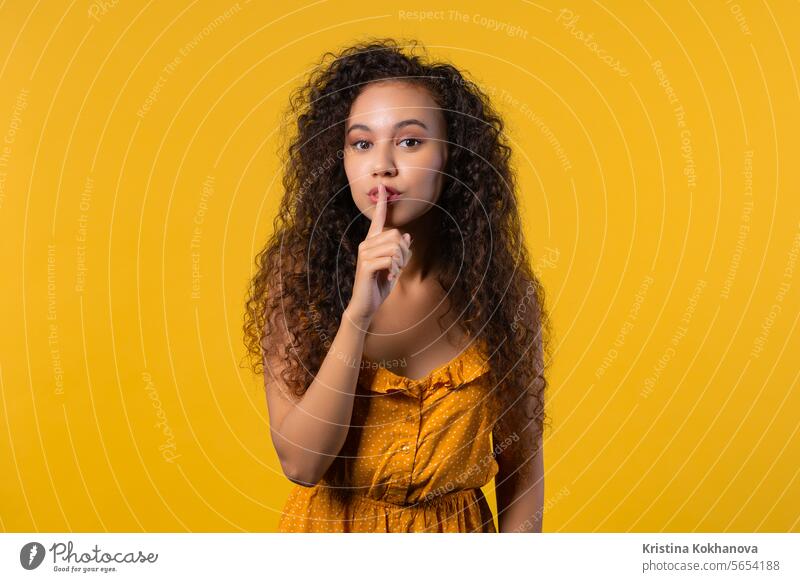 Lächelnde lockige Frau mit Finger auf den Lippen - shhh, Geheimnis, Stille, gelbes Studio fragen Hintergrund beißend charmant Nahaufnahme Konzept Verschwörung