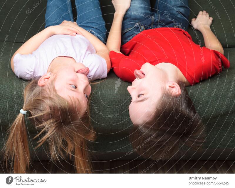 Bruder und Schwester in Freizeitkleidung liegen auf einem grünen Sofa zu Hause und strecken ihre Zungen aus, um sich gegenseitig zu necken Halbgeschwister