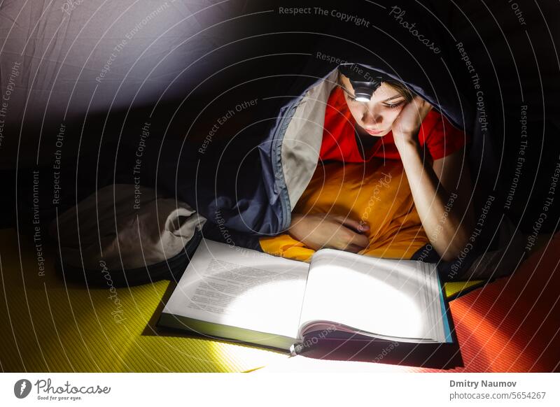 Teenager, der in einem Campingzelt liegt, eingewickelt in einen Schlafsack, und nachts mit einer Taschenlampe ein Buch liest Bett Schlafenszeit Junge Windstille