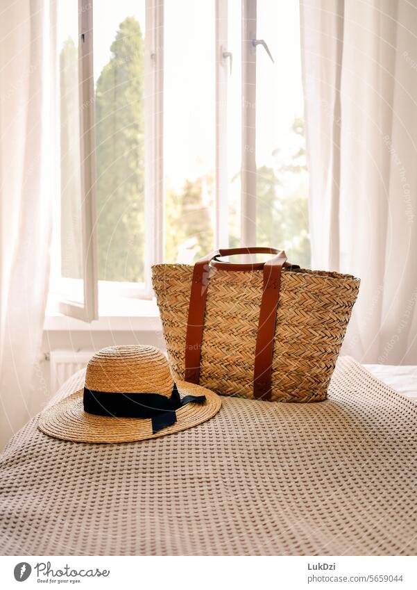 Strohhut und Korbtasche auf einem Hotelbett mit einem offenen Fenster im Hintergrund an einem sonnigen, menschenleeren Sommertag Idylle Tourismus Küste Hut