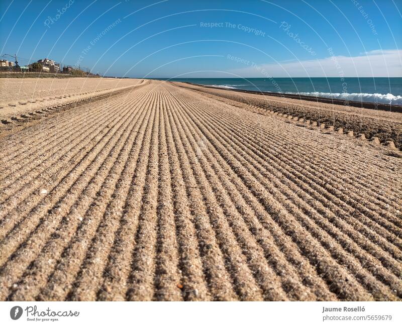 Strand von Vilassar de Mar, Barcelona. Mit vielen langen geraden Linien, die im Horizont verschwinden (die Linien sind die Spuren des Traktors, der den Sand reinigt), auf der rechten Seite das Mittelmeer, Tag mit blauem Himmel, gleich am Morgen.