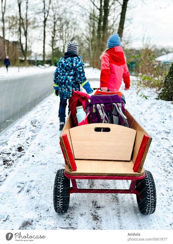 Schulweg im Winter | Zwei Kinder ziehen einen Bollerwagen mit Schulranzen Schüler Schülerin Schulkind nach hause Heimweg Schule Bildung Kindheit Schnee Straße