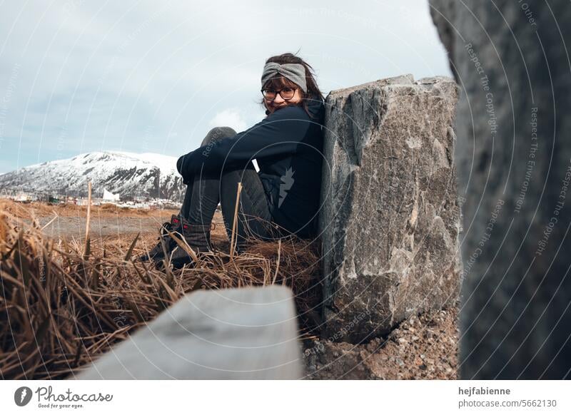 Am Polarkreis: Portrait einer jungen Frau in Winterkleidung und Stirnband, auf dem Boden sitzend und mit angewinkelten Beinen an einen Stein gelehnt Outdoor