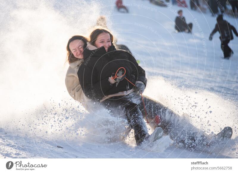 Zwei Jugendliche rasen schneestaubend auf einem Schlitten durch den Sonnenschein Schnee Winter schlittenfahren Kinder Sonnenlicht Rodeln Rodelbahn kalt Piste