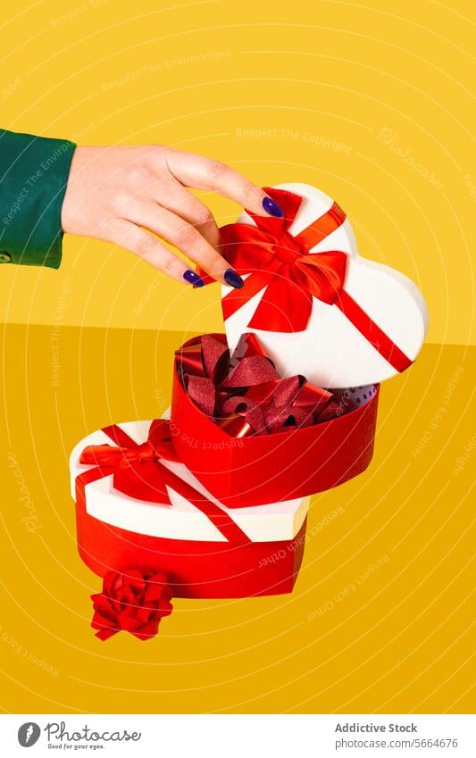 Anonyme Hand, die eine herzförmige Geschenkbox mit roten Bändern auf gelbem Hintergrund öffnet Kasten Herz Form Bändchen Eröffnung präsentieren Valentinsgruß