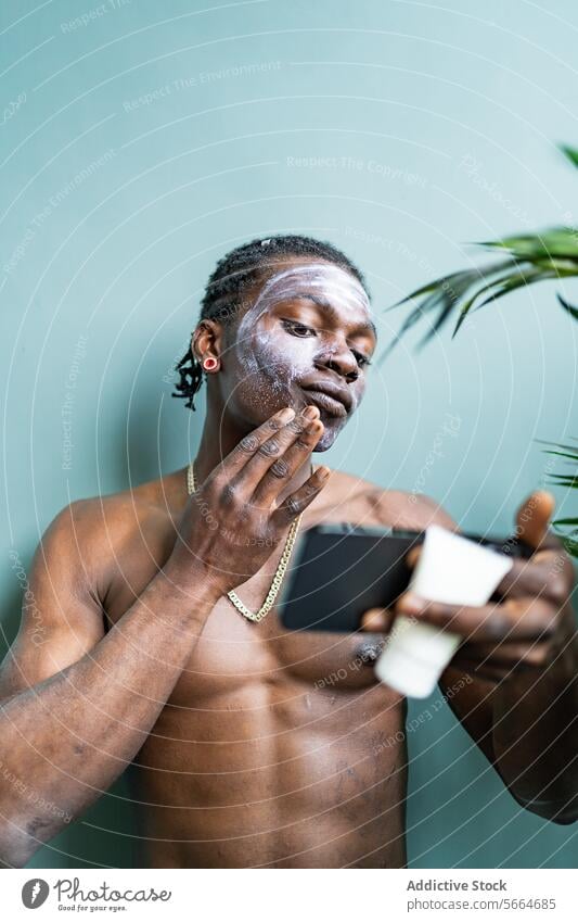 Schwarzer Mann wendet Gesichtsmaske an, während er sein Spiegelbild auf einem Telefon vor einem weichen grünen Hintergrund überprüft Hautpflege
