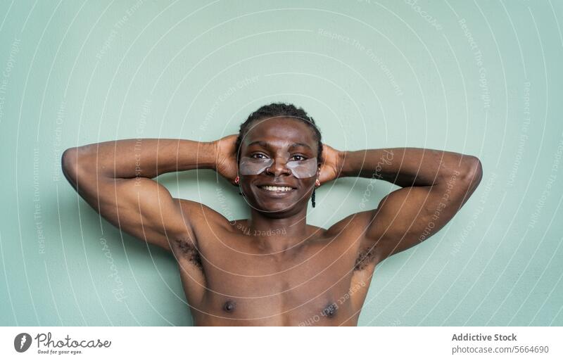 Ein fröhlicher schwarzer Mann mit Augenklappen lächelt in die Kamera, die Hände hinter dem Kopf vor einer mintgrünen Wand. Hautpflege Lächeln Freude