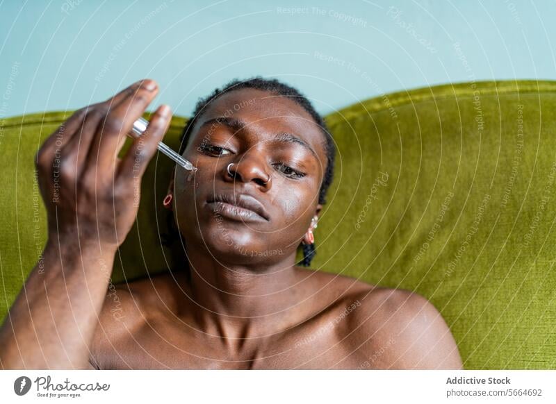 Ein schwarzer Mann trägt ein Serum mit einer Pipette auf sein Gesicht auf, während er auf einem grünen Sofa sitzt und sich intensiv mit seiner Hautpflege beschäftigt