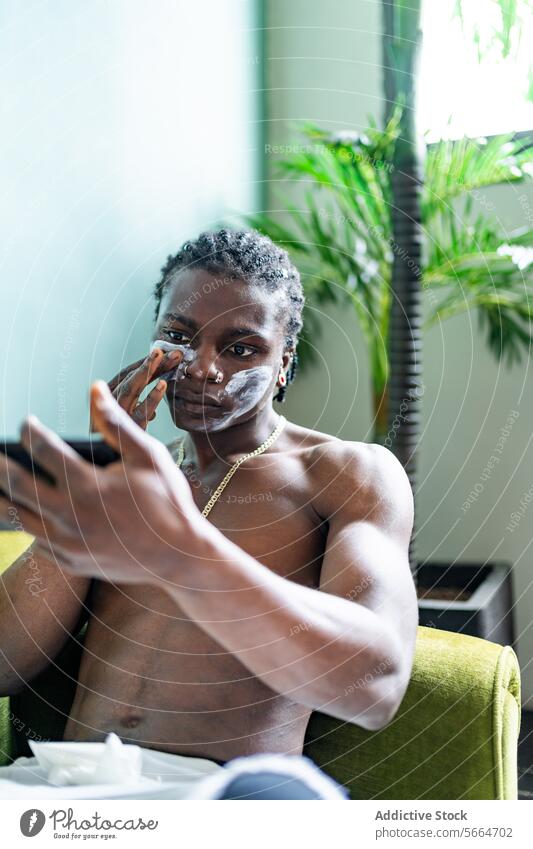 Ein fokussierter schwarzer Mann trägt eine Gesichtsmaske auf, während er sein Spiegelbild auf einem Telefon betrachtet, das in einem Haus mit Pflanzen im Hintergrund steht.