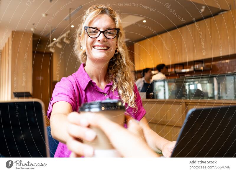Kaukasische, fröhliche, blonde Frau mit Brille, die eine Kaffeetasse in der Hand hält und einen Moment mit einem Kollegen in einem Café während der Fernarbeit in Chiang Mai, Thailand, teilt