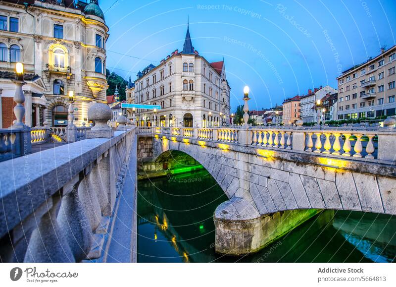 Die Dämmerung legt sich über die kopfsteingepflasterten Straßen von Ljubljana, die Dreifachbrücke ist beleuchtet und die Gebäude spiegeln sich im Fluss Ljubljanica