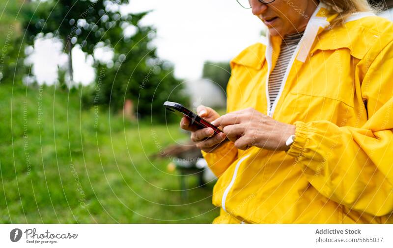 Fokussierter Ausschnitt einer Imkerin in gelbem Overall, die ein Mobiltelefon benutzt, während sie im grünen Gras steht Frau Smartphone Bienenkorb gesamt Handy