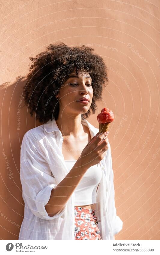 Eine Frau mit lockigem Haar genießt eine Erdbeereiswaffel vor einer pfirsichfarbenen Wand, die eine entspannte Sommeratmosphäre verkörpert krause Haare
