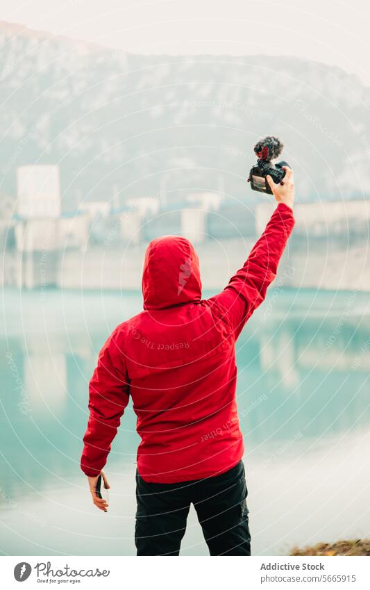 Rückenansicht Person in einem roten Kapuzenpulli, die ein Selfie mit einer Kamera vor einer ruhigen See- und Dammlandschaft macht roter Kapuzenpulli Fotokamera