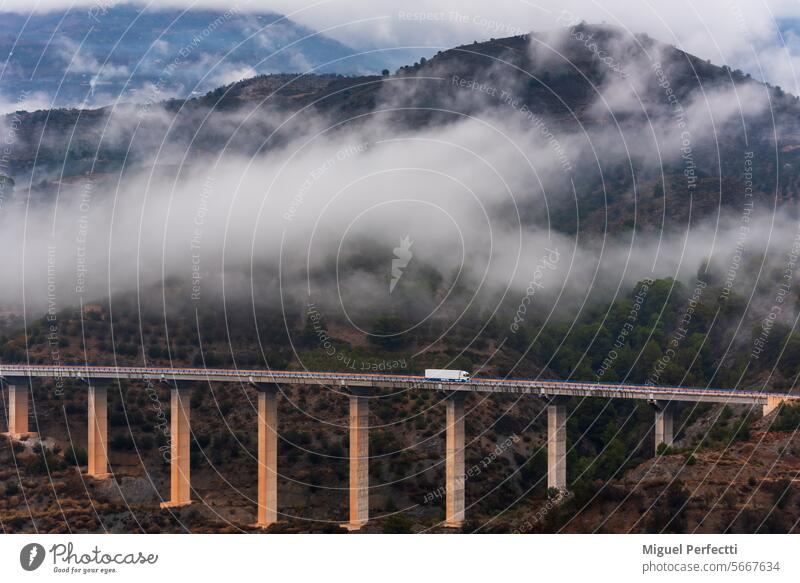Lastwagen mit Kühlsattelauflieger, der über eine Brücke und eine Berglandschaft mit tief hängenden Wolken im Hintergrund fährt. Verkehr Berge u. Gebirge