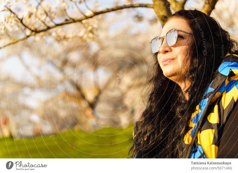 Frau mit Sonnenbrille mit verspiegelten Gläsern und langen dunklen Haaren im blühenden Frühlingsgarten in einem öffentlichen Park an einem sonnigen Frühlingstag