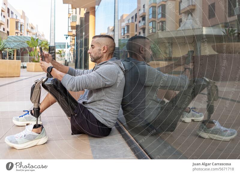 Männlicher Amputierter benutzt Smartphone in der Nähe einer Glaswand Sportler benutzend Training Pause Prothesen Fitness online männlich reif Lebensmitte sitzen