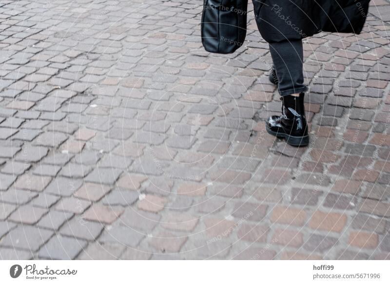 Teilaufnahme eines auf Kopfsteinpflaster gehenden, schwarz gekleideten Menschen von schräg hinten Weg schwarze Kleidung Person Straße urban Bekleidung Großstadt