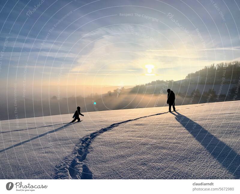 Ein Kind und ein Erwachsener im Schnee, bei Sonnenuntergang und Gegenlicht. Das Kind geht auf den Erwachsenen zu. Schneelandschaft Winter Sonnenlicht Wege