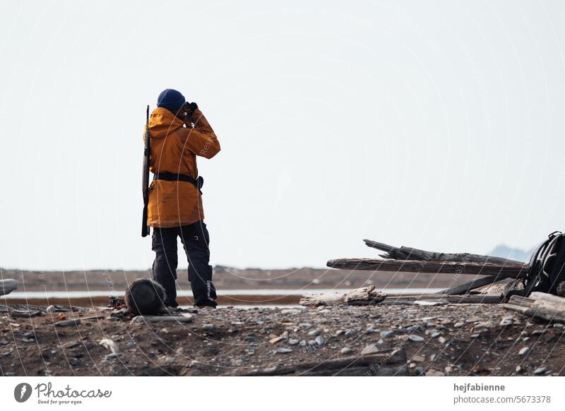 Eisbärwache auf Spitzbergen während einer Expedition. Weiblicher Guide mit gelber Ranger-Jacke und Gewehr in arktischer Tundra überwacht die Umgebung mit einem Fernglas.