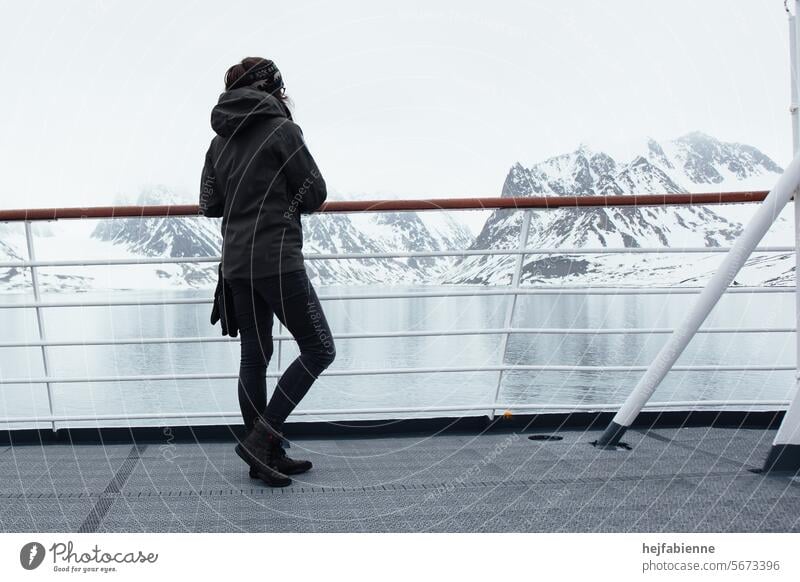 Eine junge Frau mit Winterkleidung steht an der Reling eines Kreuzfahrtschiffs und blickt auf einen arktischen Fjordw während einer Spitzbergenexpedition.