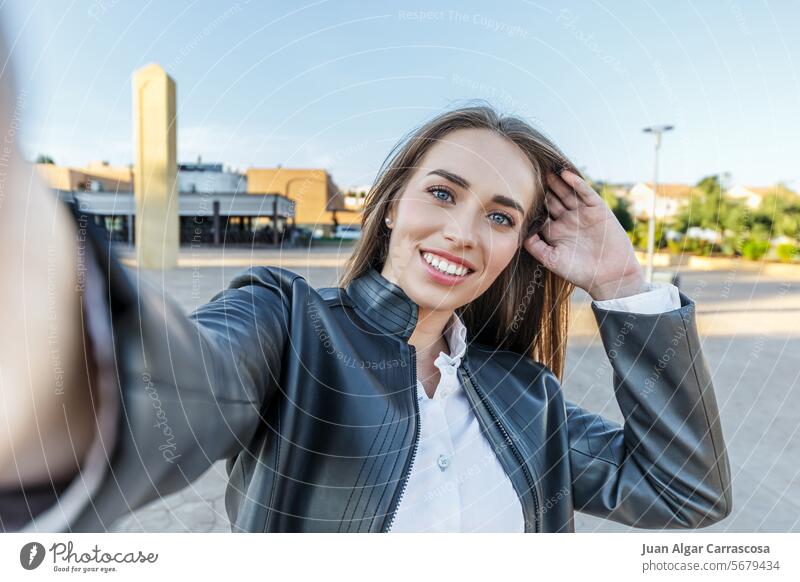 Blauäugige blonde Frau in Lederjacke macht lächelnd ein Selfie mit ihrer Kamera auf der Straße Porträt ihr Haar zu berühren Fröhlichkeit Frisur Lächeln