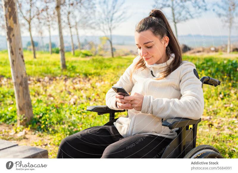 Eine junge Frau im Rollstuhl lächelt, während sie in einem Park auf ihrem Smartphone surft Lächeln Bank Handy Browsen im Freien Freizeitkleidung Sitzen