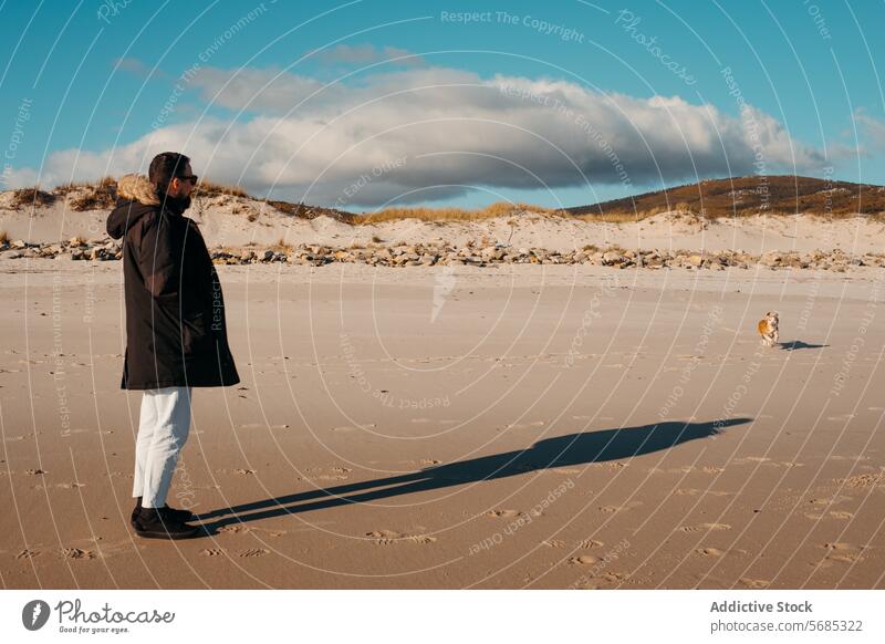 Mann im Mantel steht an einem Sandstrand und sieht einen Hund in der Ferne, mit Dünen und blauem Himmel über ihm in Lariño, Galicien Strand Entfernung Galicia