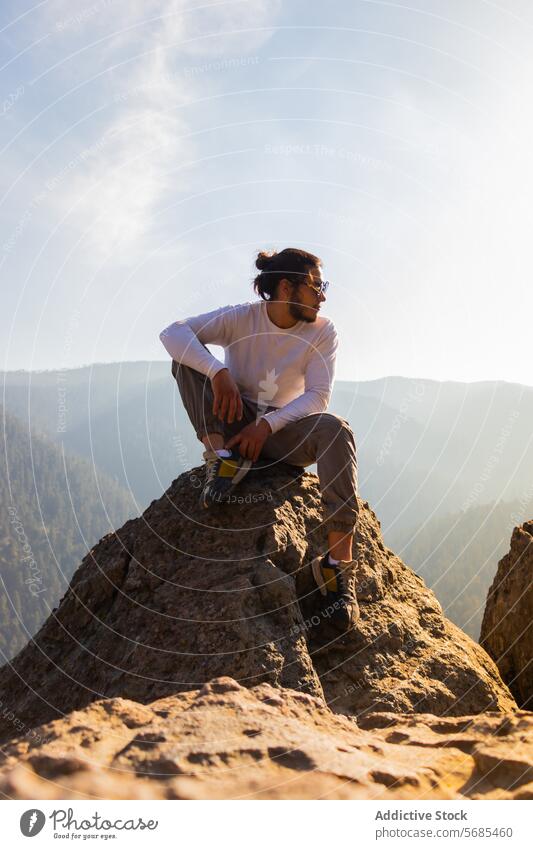 Junger Mann sitzt auf einer Klippe in der Nähe der nebligen Berge sitzen Berge u. Gebirge bewundern Tourist lässig Outfit Vollbart Sonnenbrille felsig Natur