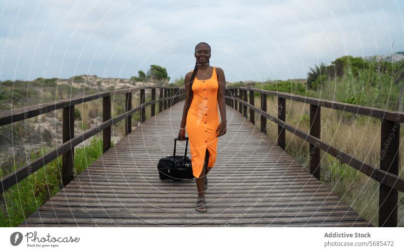 Lächelnde schwarze Frau, die auf einem Holzsteg geht Spaziergang hölzern Steg Dame Kleid Ziehen Gepäck positiv Reisender Hügel Berge u. Gebirge Natur reisen